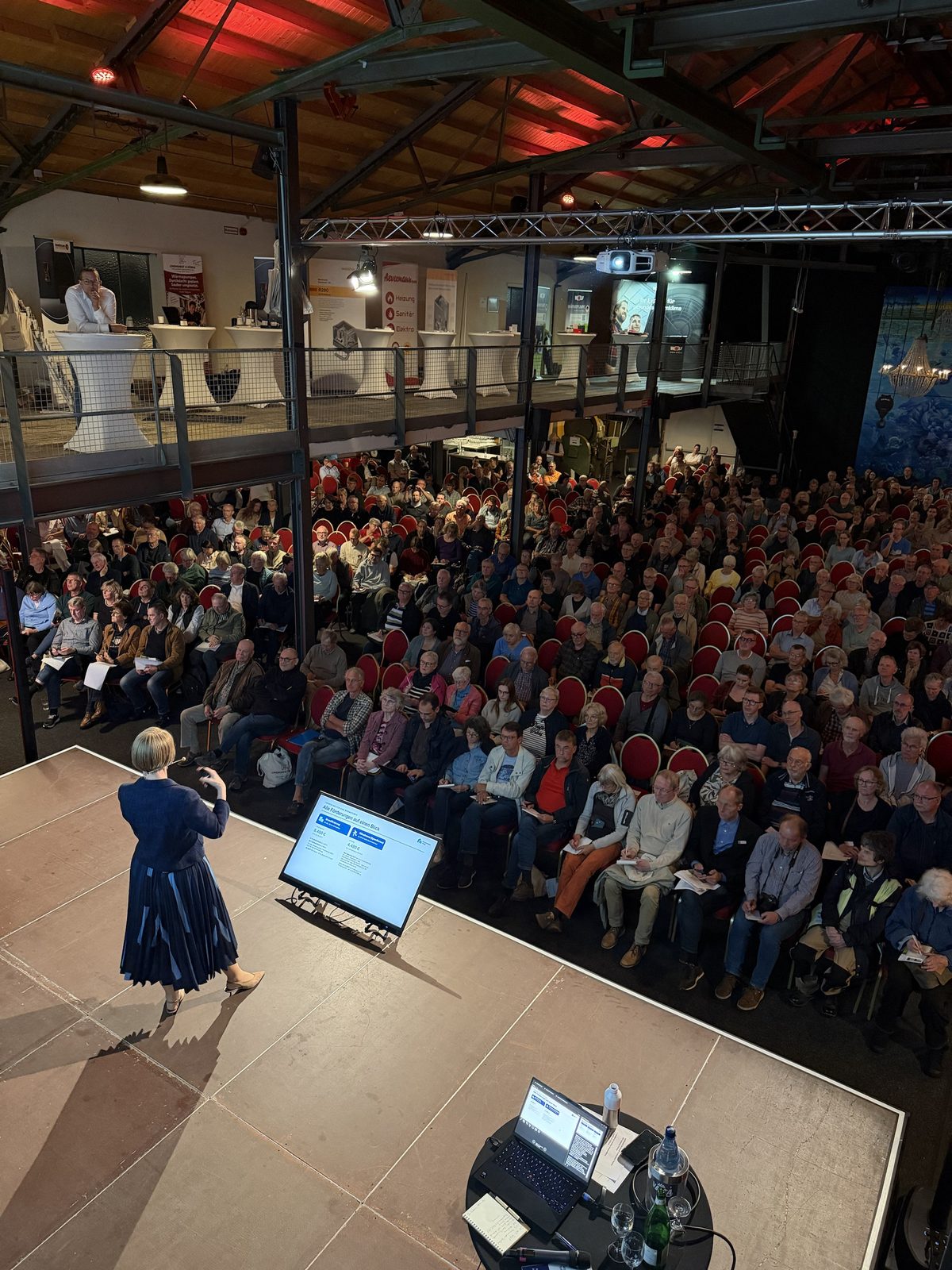 Anja Floetenmeyer-Woltmann im Vortrag zur Förderung, vor ihr die vollen Stuhlreihen, oben auf der Galerie die Stände der Fachbetriebe