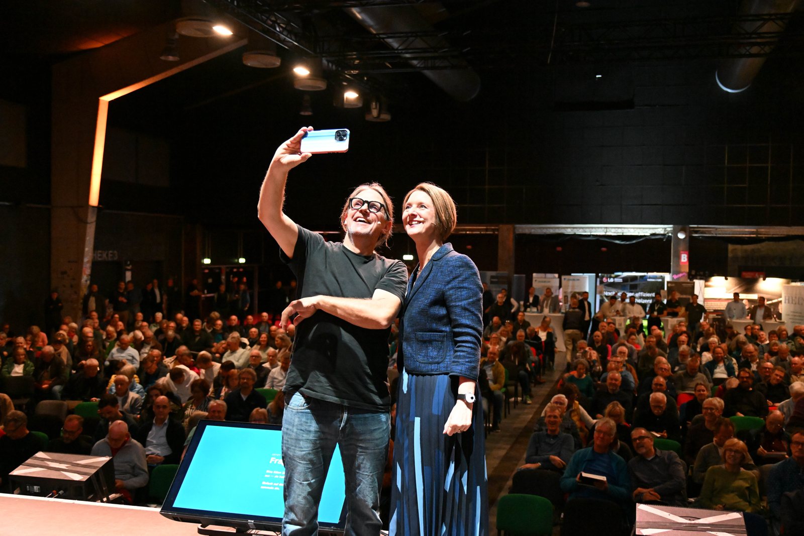 Carsten Herbert und Anja Floetenmeyer-Woltmann machen ein Selfie auf der Bühne vor dem vollen Saal